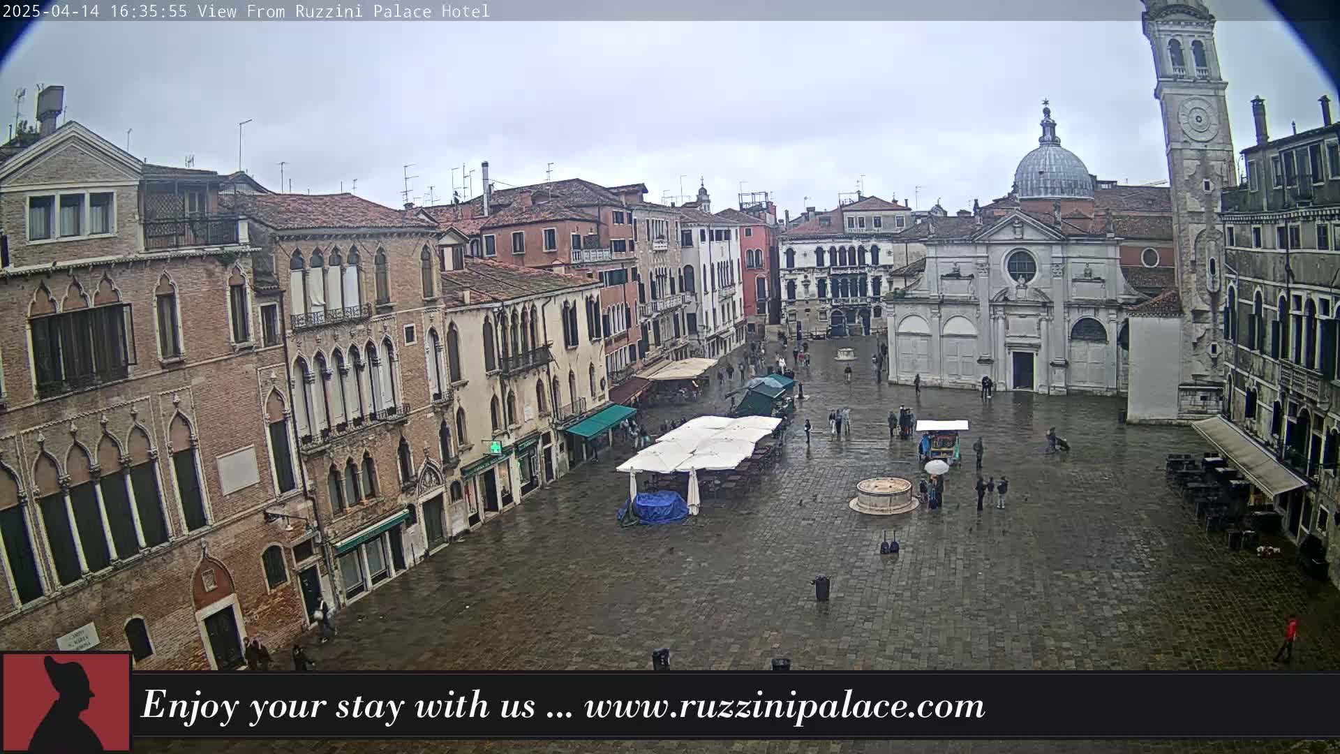 A wet, cobblestone square in Venice, Italy, is surrounded by old, multi-story buildings, one of which has a dome and tall clock tower.