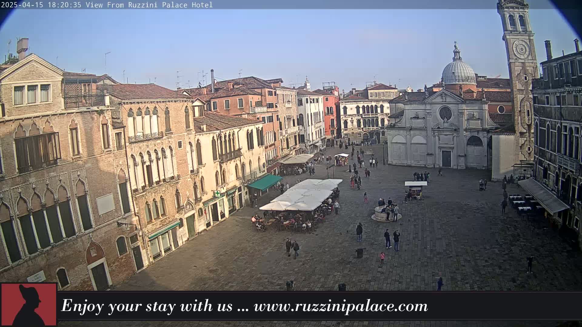 A mostly sunny view of a large, paved town square in Venice, Italy, with several buildings surrounding it, people walking around, and outdoor seating under umbrellas.