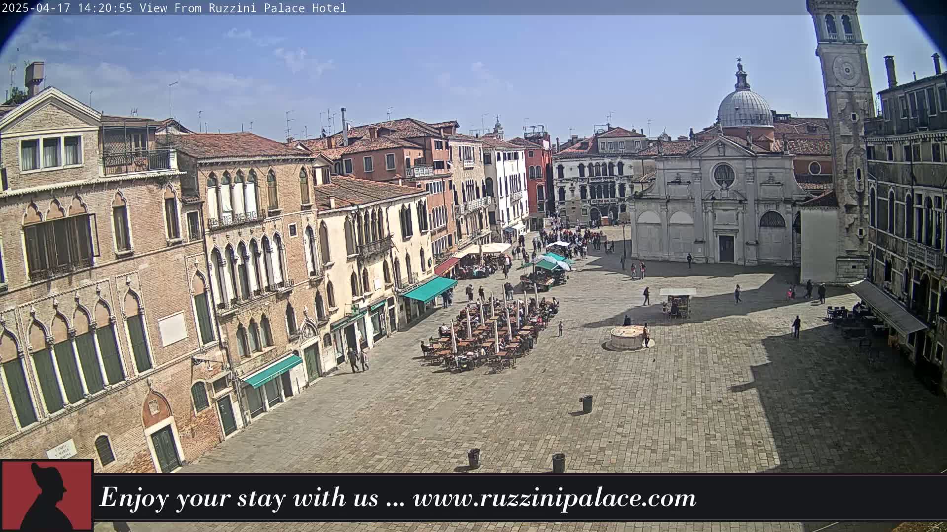 A sunny Venetian plaza is shown with several buildings surrounding it, an outdoor cafe with patrons, and a church with a dome and bell tower.