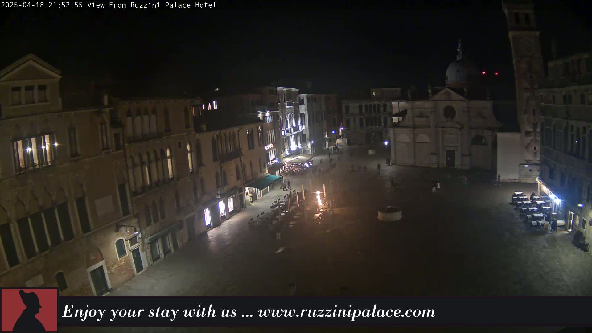 A nighttime, high-angle view of a dimly lit plaza in Venice, Italy, featuring several buildings, outdoor seating areas, and a church.