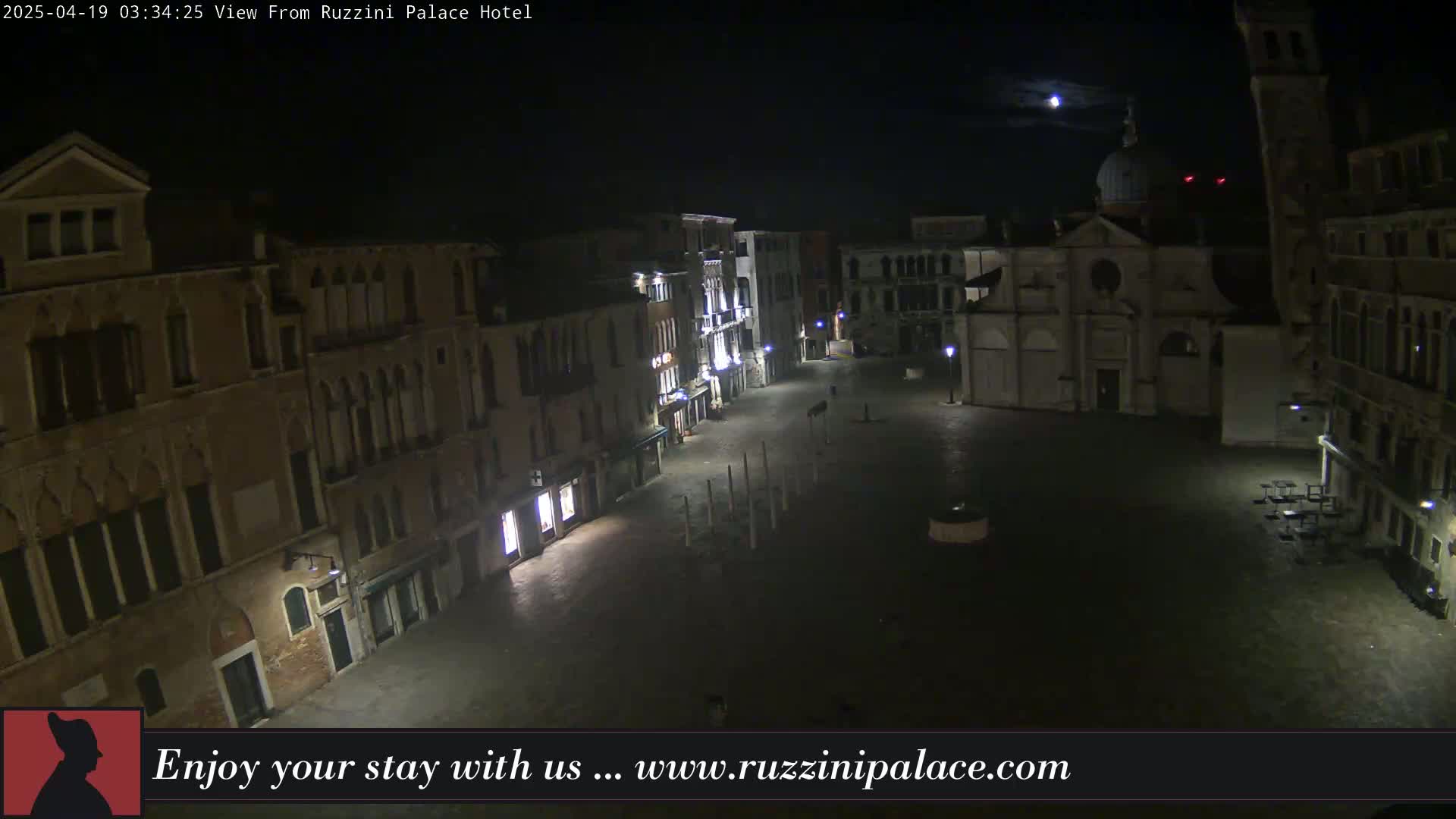 A nighttime view of a mostly empty plaza in Venice, Italy, under a partly cloudy moonlit sky, shows several historic buildings surrounding the square.
