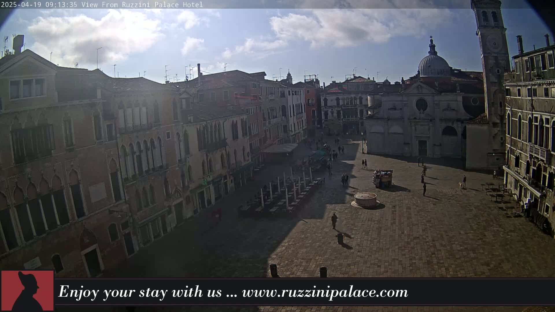 A mostly sunny day reveals a Venetian square with several buildings, a few people walking around, and outdoor seating.