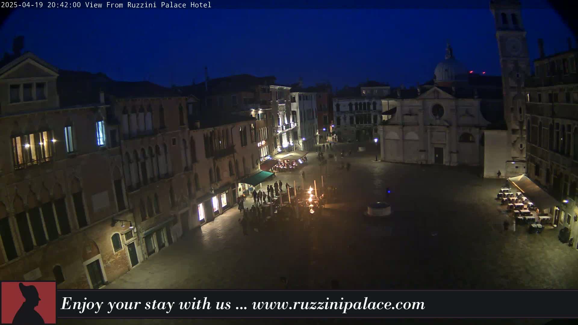 A nighttime aerial view of a dimly lit European town square, surrounded by old buildings, with a few people and some outdoor seating areas visible.