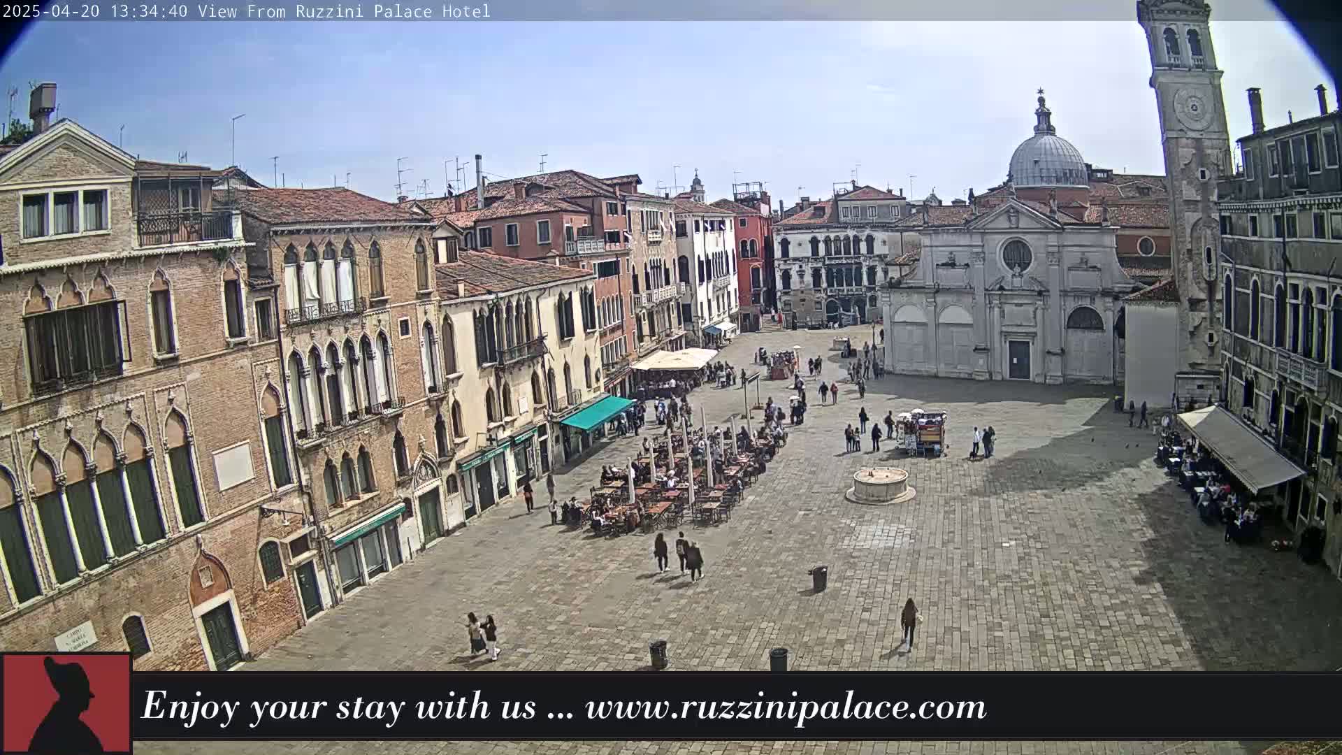 A sunny day in a large, paved Venetian square with several buildings, a church, and people sitting at outdoor cafes.