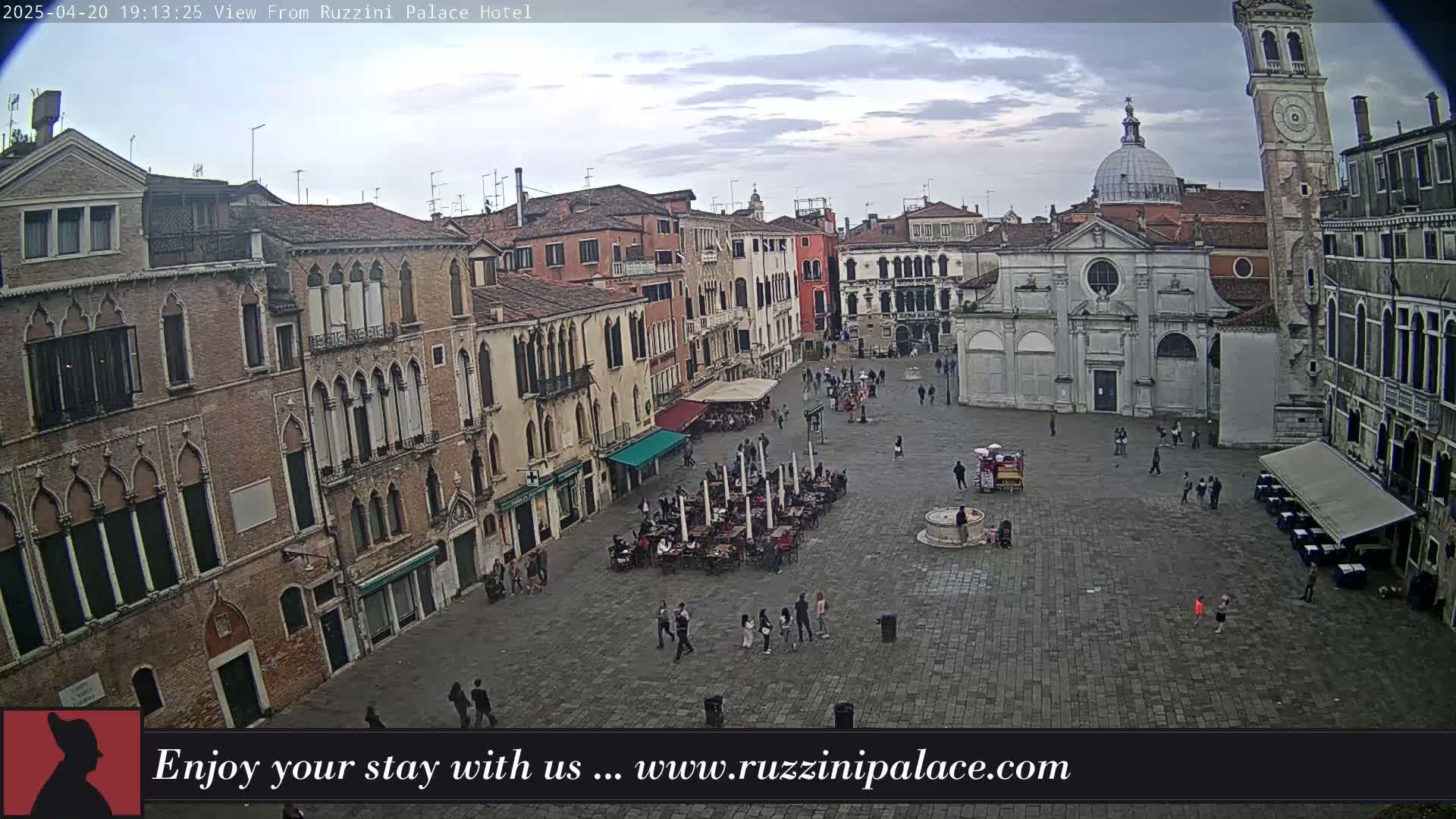 An overcast sky hangs over a large, stone plaza in Venice, Italy, surrounded by historic buildings and populated by a scattering of people sitting at outdoor cafes and walking.
