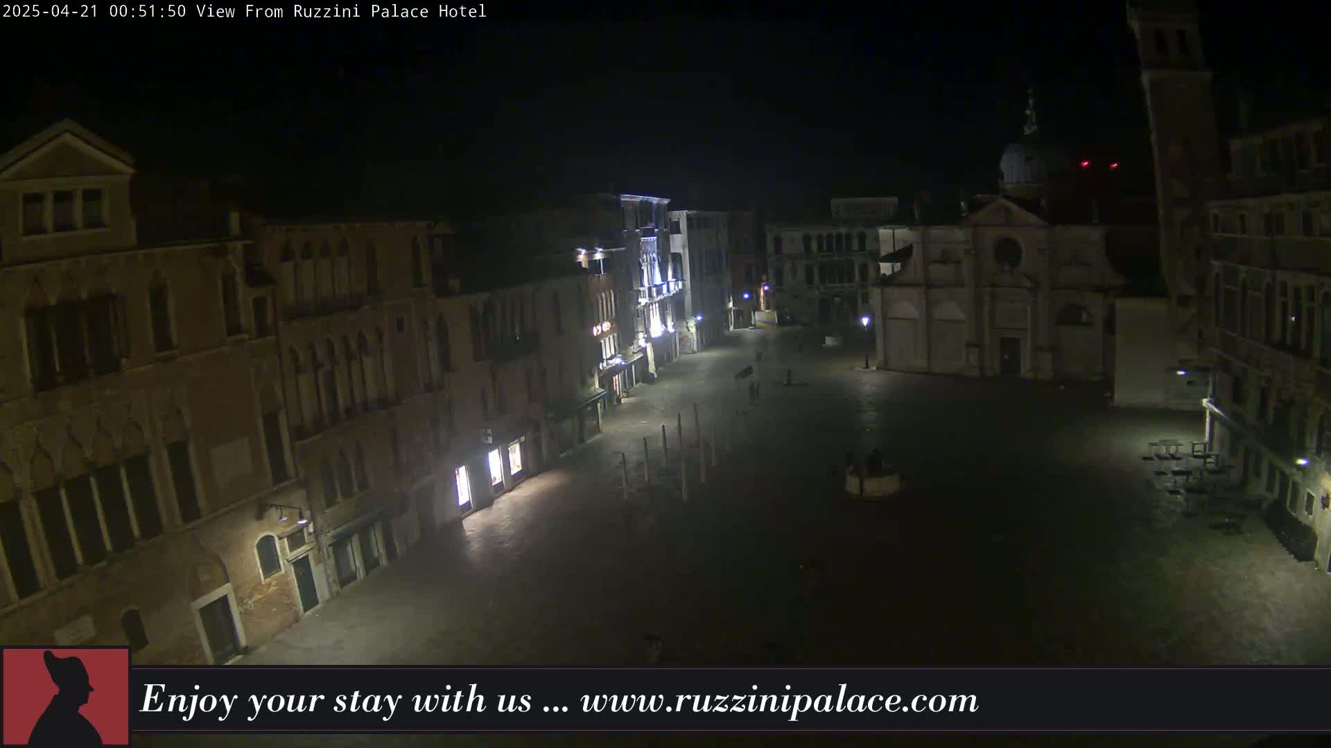 A nighttime, high-angle view of a mostly empty plaza in Venice, Italy, surrounded by old buildings.