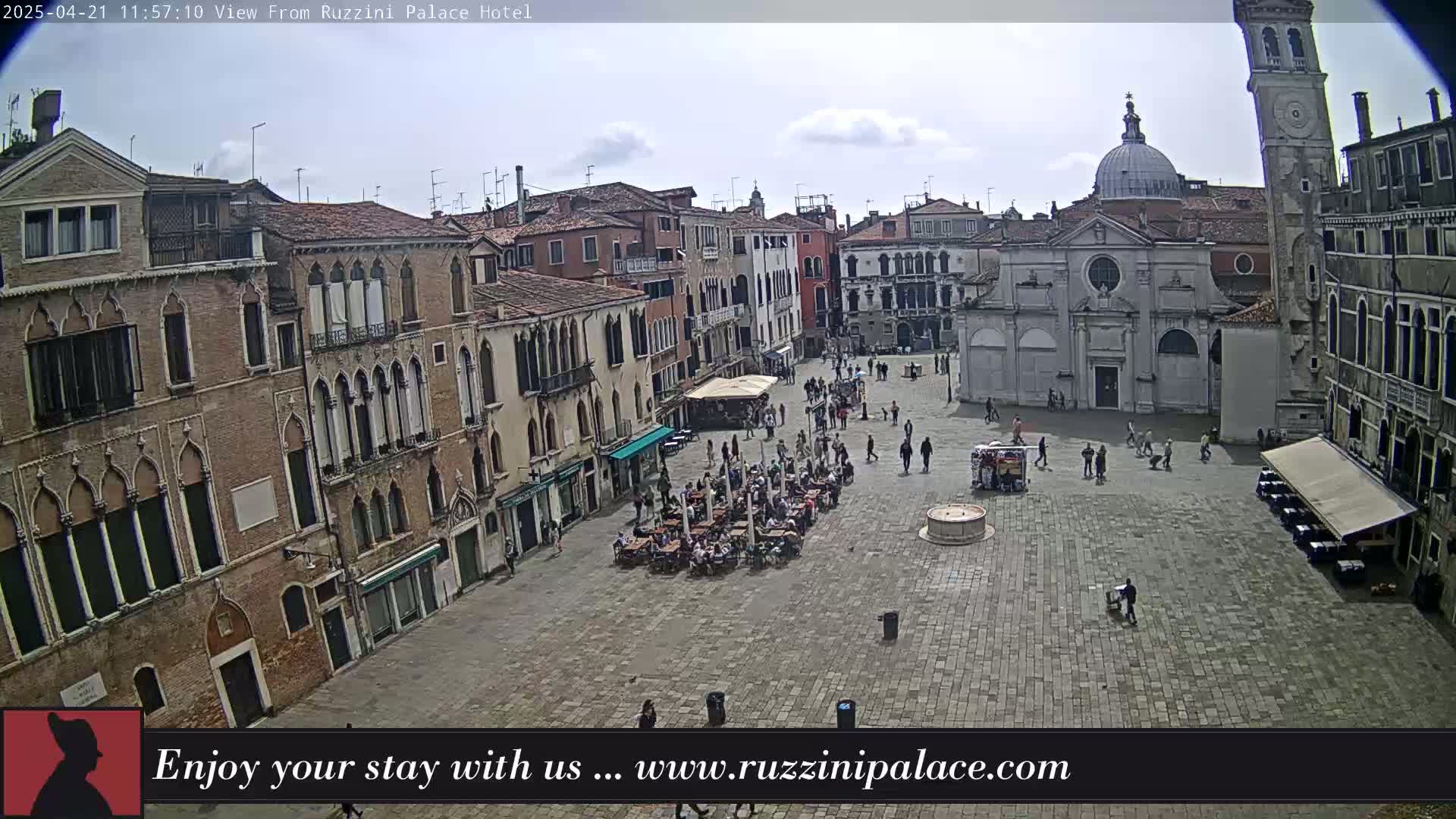 A mostly sunny day shows a large cobblestone square in Venice, Italy, filled with people and outdoor seating, surrounded by old brick buildings, including a church and tall tower.