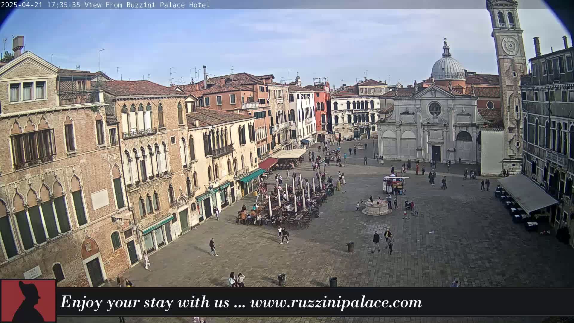 A sunny outdoor view shows a large town square in Venice, Italy, filled with people and surrounded by historic buildings, including a church and a leaning tower.
