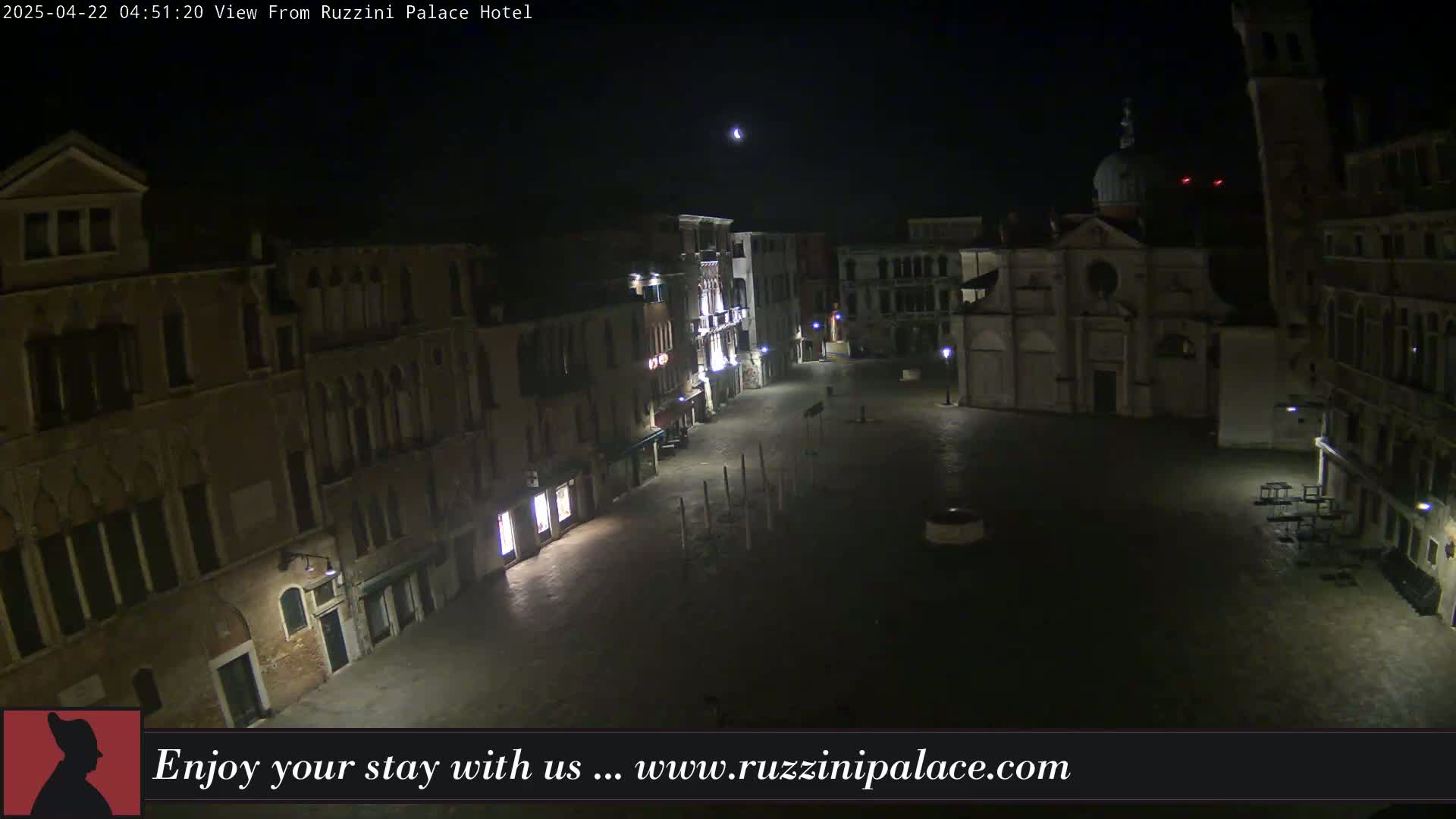A nighttime, high-angle view of a dimly lit, empty European-style square surrounded by old buildings, under a clear night sky with a visible moon.