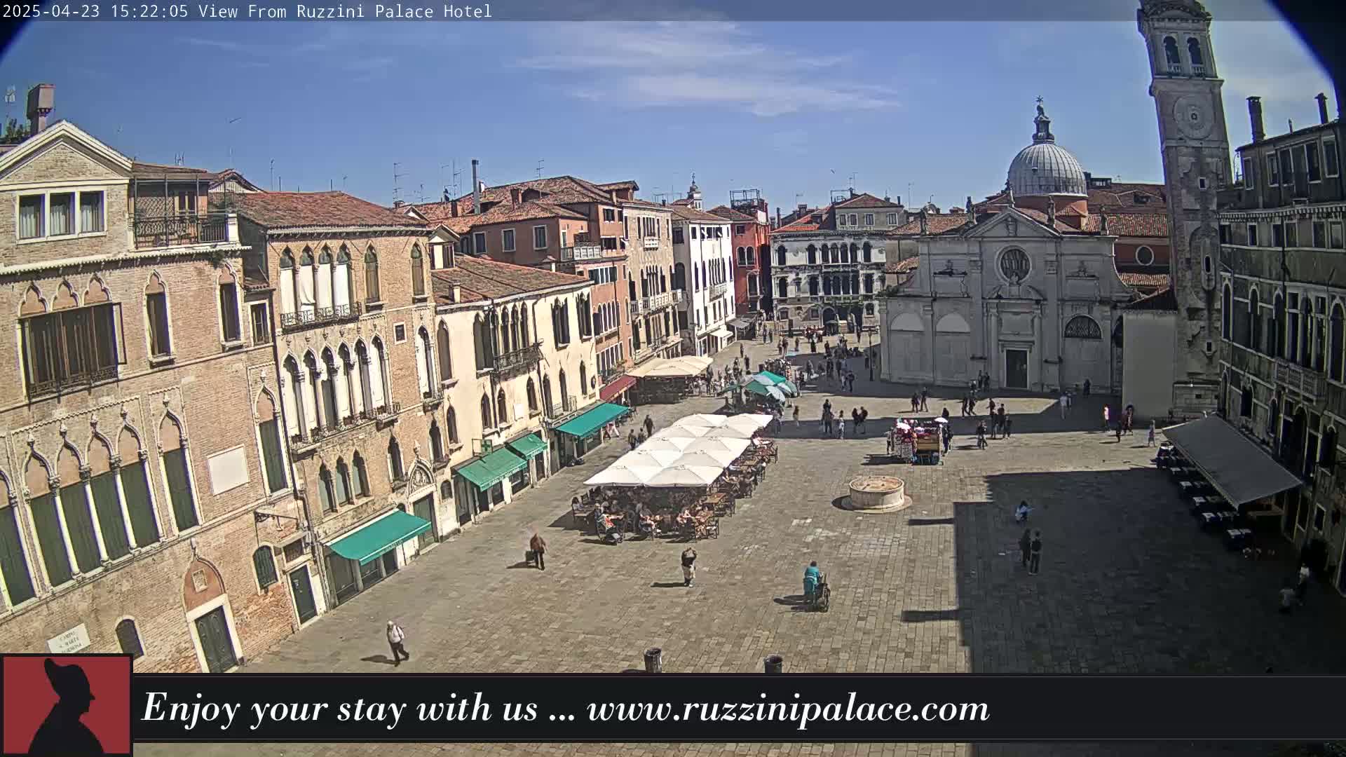 A sunny day view of a large plaza in Venice, Italy, filled with people and outdoor seating, surrounded by historic buildings.