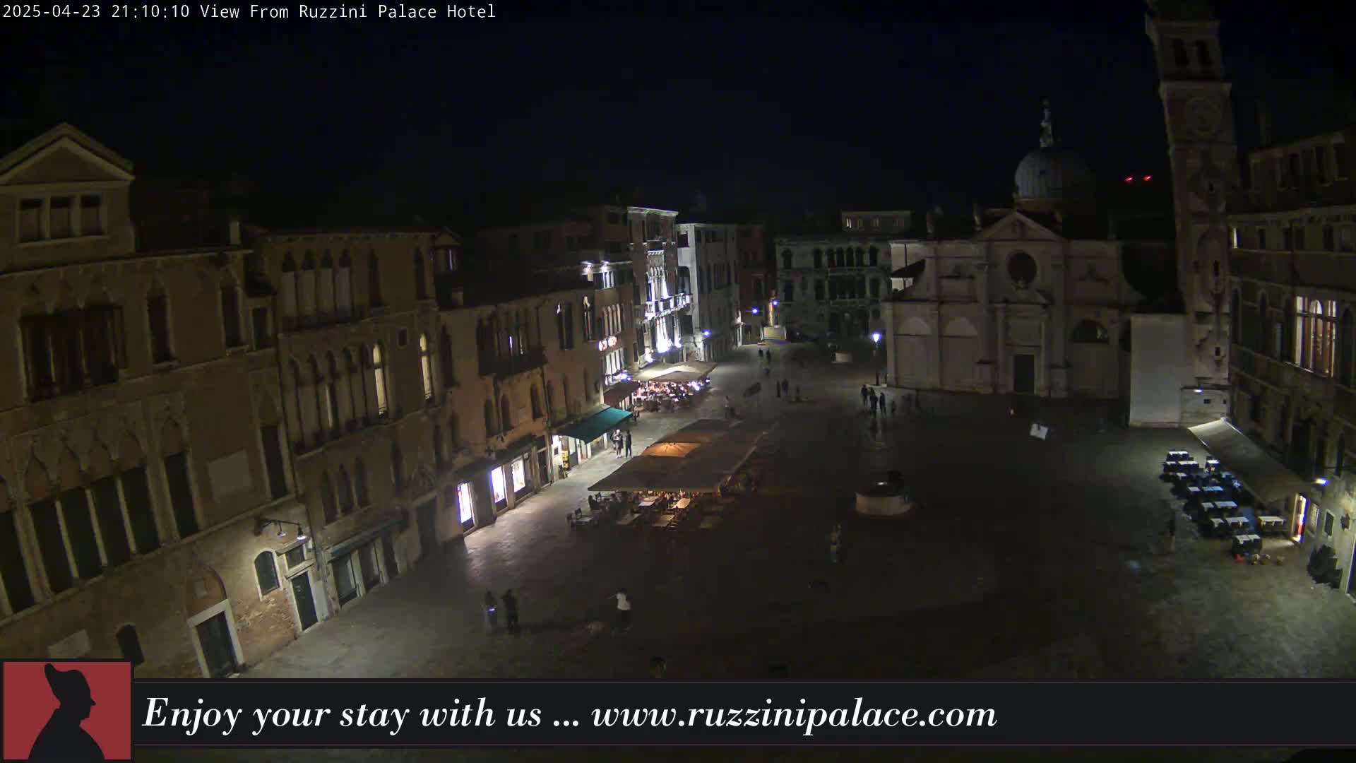 A nighttime view of a dimly lit European-style square with several buildings, a church, and a few people walking around.