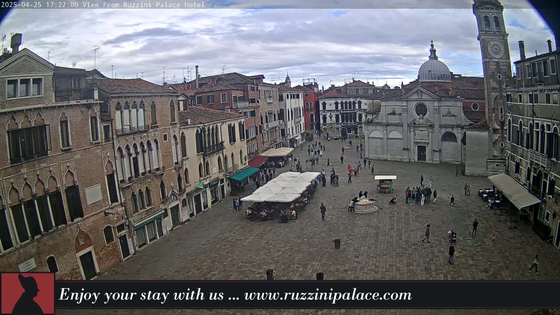 An overcast day reveals a large, stone plaza in Venice, surrounded by historic buildings, with people strolling and several outdoor cafes set up.