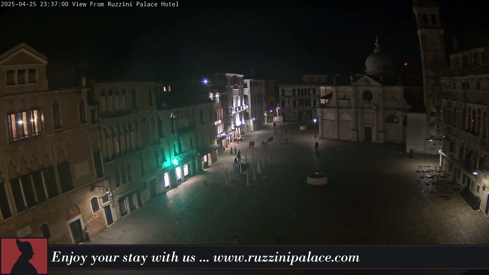 A nighttime view of a dimly lit, mostly empty plaza in Venice, surrounded by tall buildings.