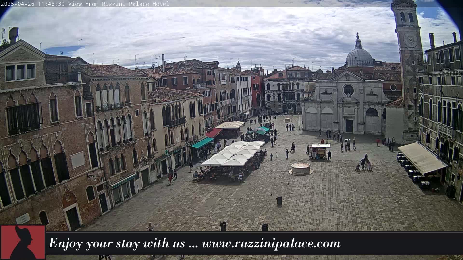 A mostly cloudy day reveals a cobblestone square in Venice, Italy, surrounded by aged buildings, with several people and outdoor café umbrellas scattered throughout.