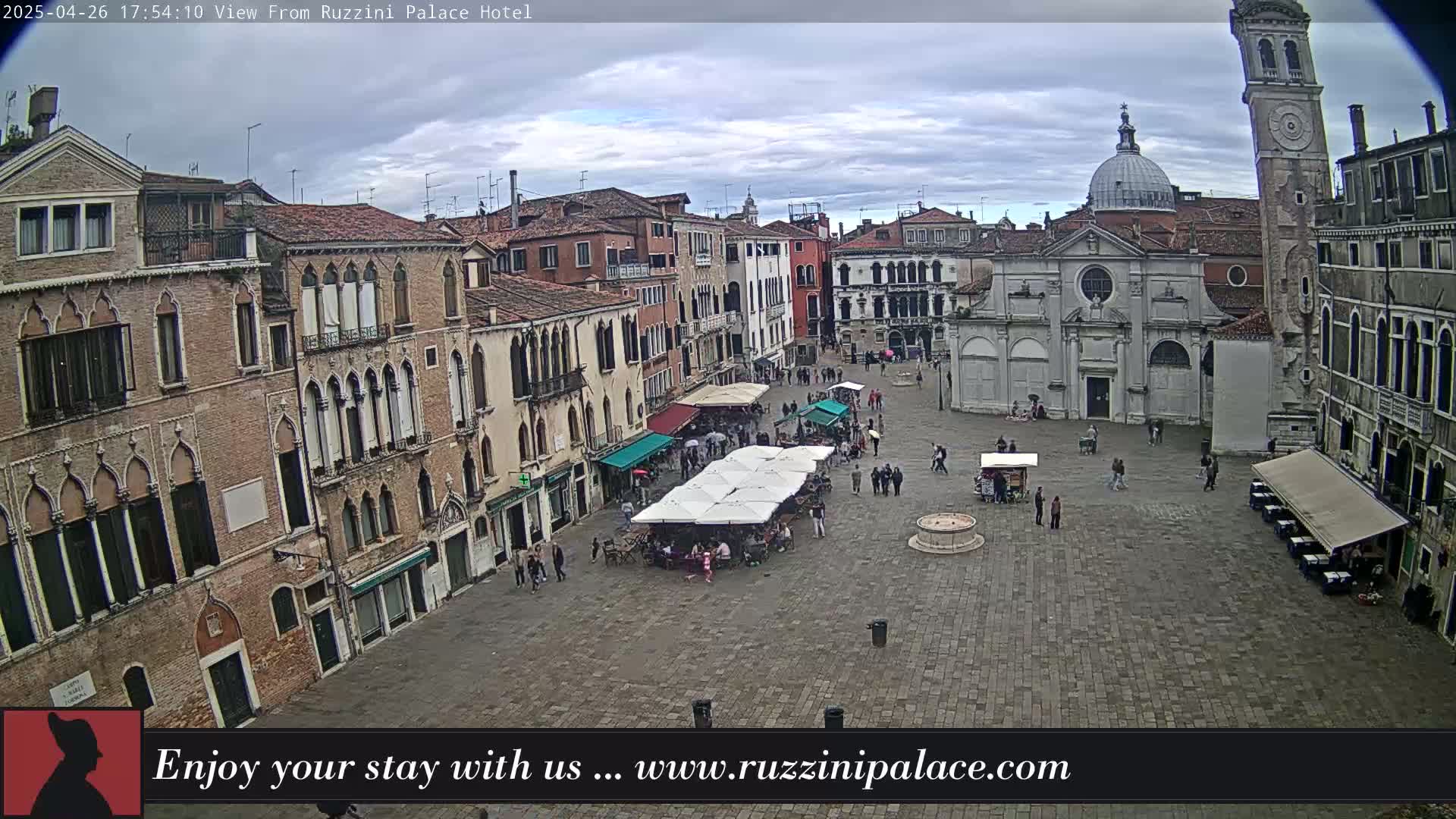 A cloudy day reveals an aerial view of a large, cobblestone square in Venice, Italy, surrounded by historic buildings and several outdoor cafes with patrons.