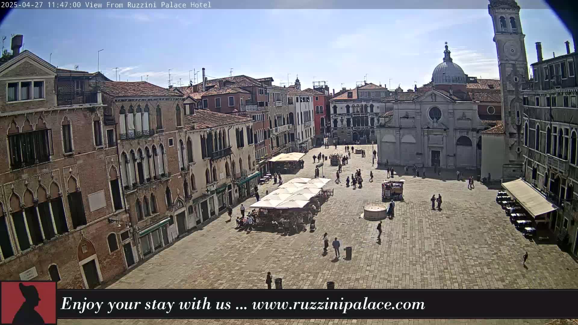 A sunny day in a large, paved square in Venice, Italy, surrounded by historic buildings, with several people walking and some seated at outdoor cafes.