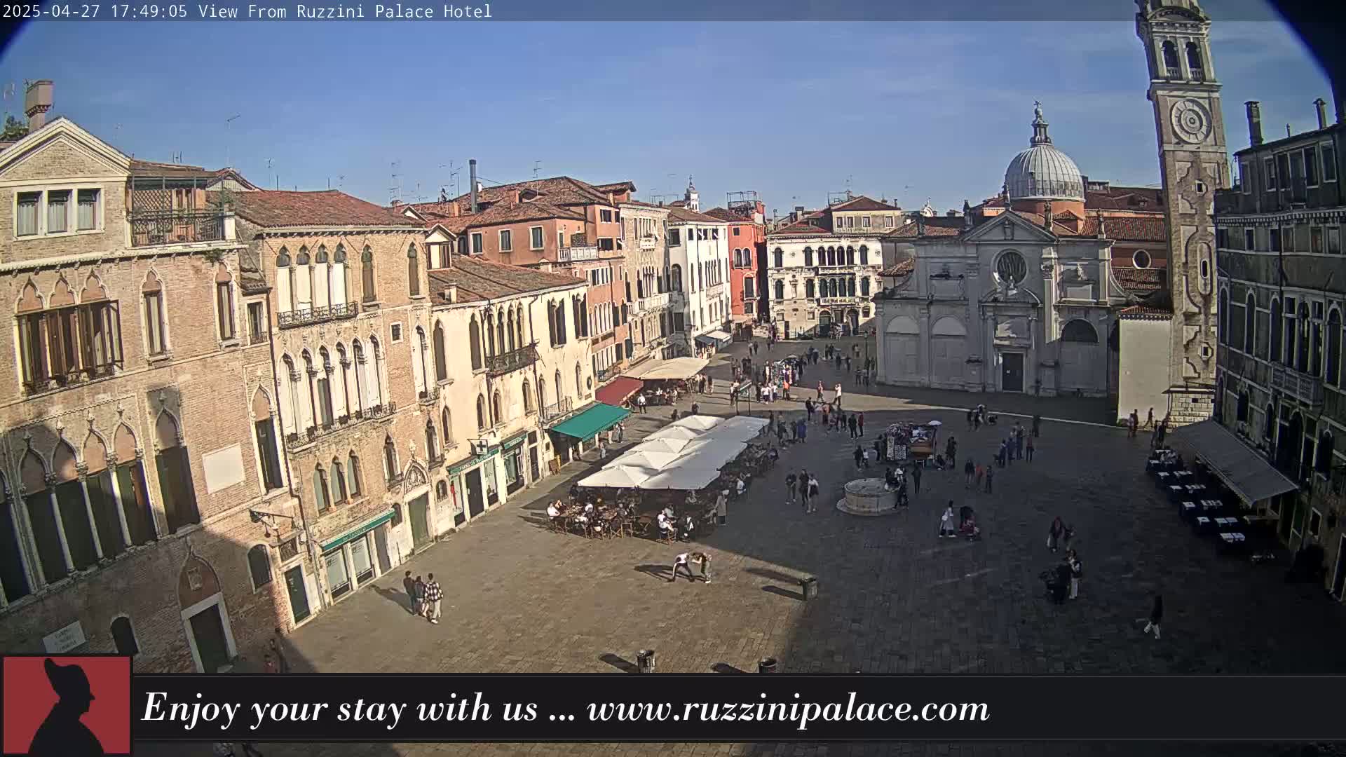 A sunny, outdoor view of a large, paved square in Venice, Italy, surrounded by historic buildings, with people walking and sitting at outdoor cafes.