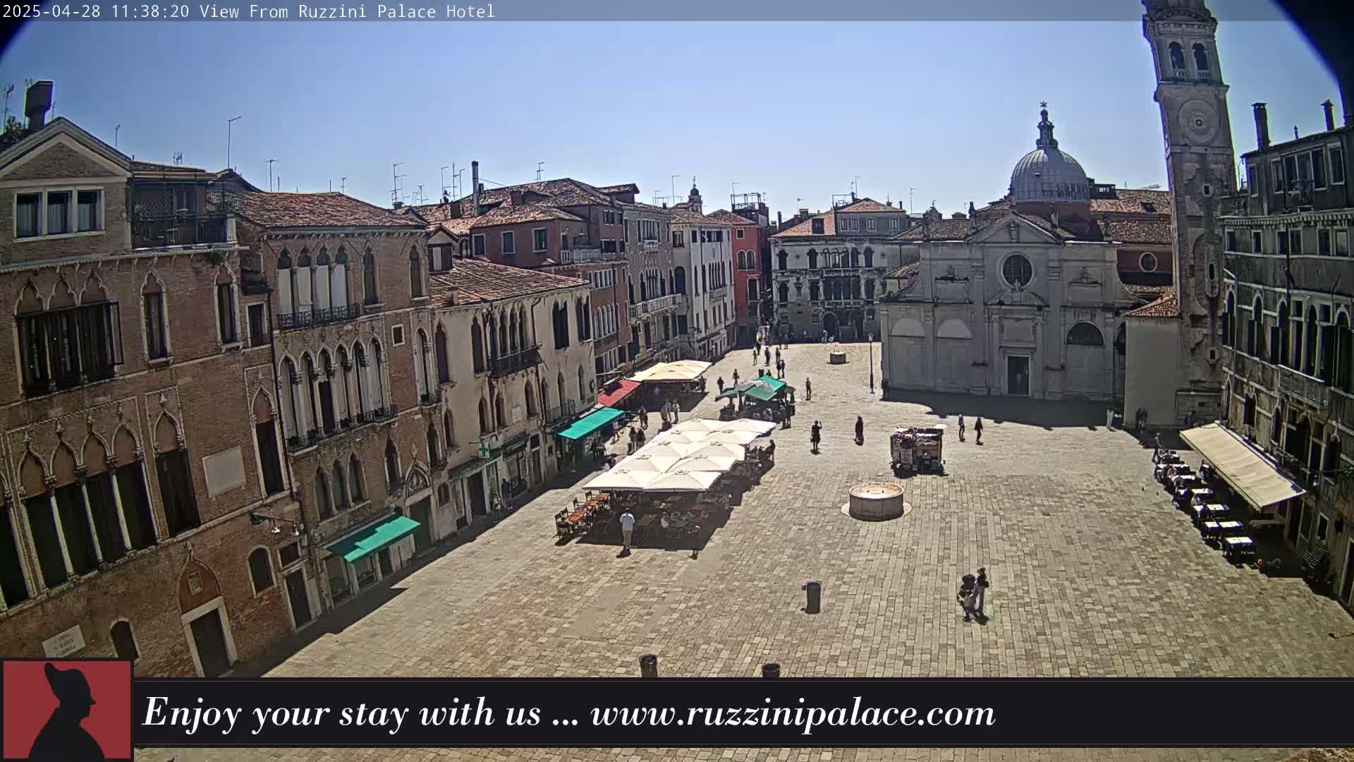 A sunny day reveals a large, paved square in Venice, Italy, surrounded by historic buildings with arched windows and outdoor cafes with umbrellas.