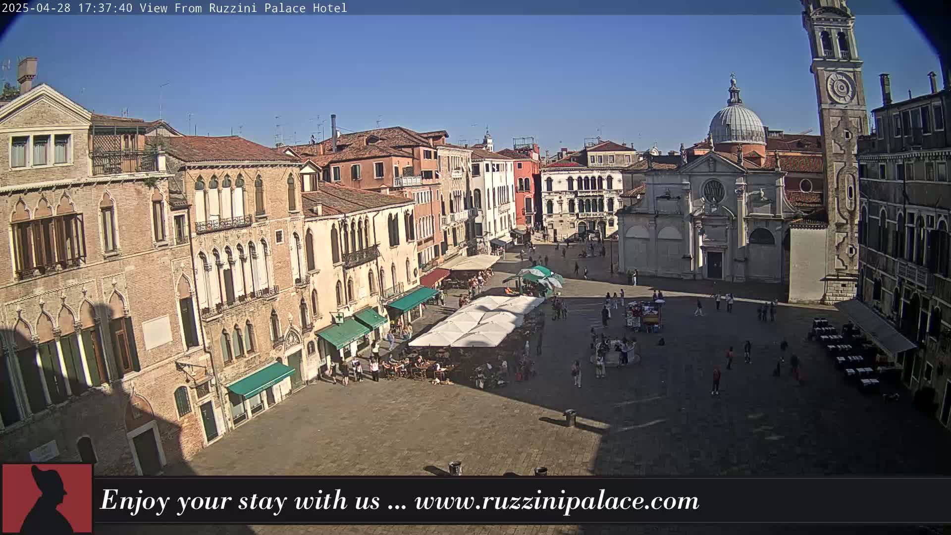 A sunny view of a large, paved plaza in Venice, Italy, surrounded by historic buildings, with people scattered throughout and several outdoor cafes with umbrellas.