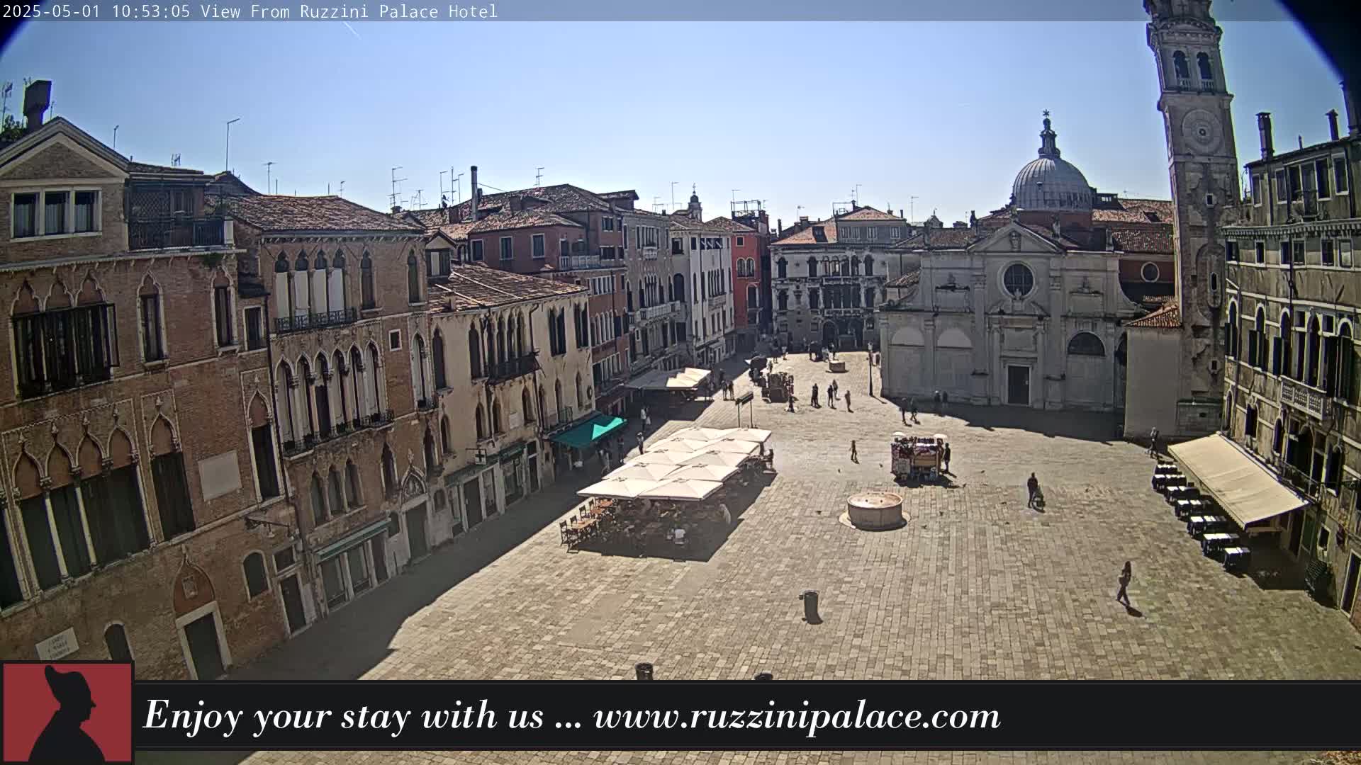 A sunny, paved square in a city is surrounded by old, multi-storied buildings, some with outdoor seating areas.