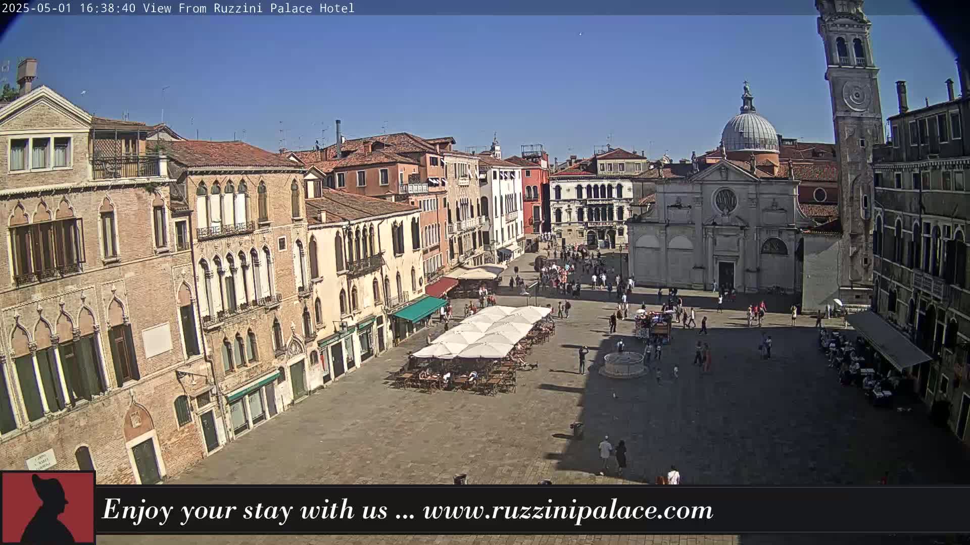 A sunny day in a large plaza in Venice, Italy, shows many people walking around and sitting at outdoor cafes amidst several historic buildings, including a church and a tall clock tower.
