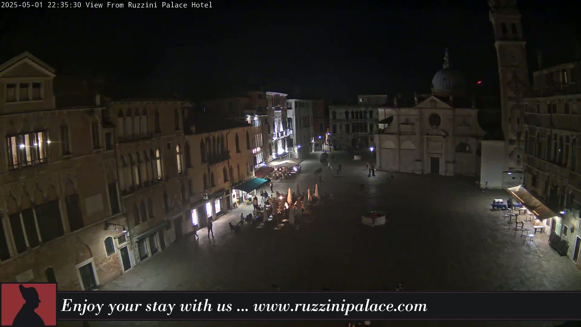 A nighttime, aerial view of a dimly lit, mostly empty town square in Venice, featuring several buildings and a few people scattered around outdoor seating areas.