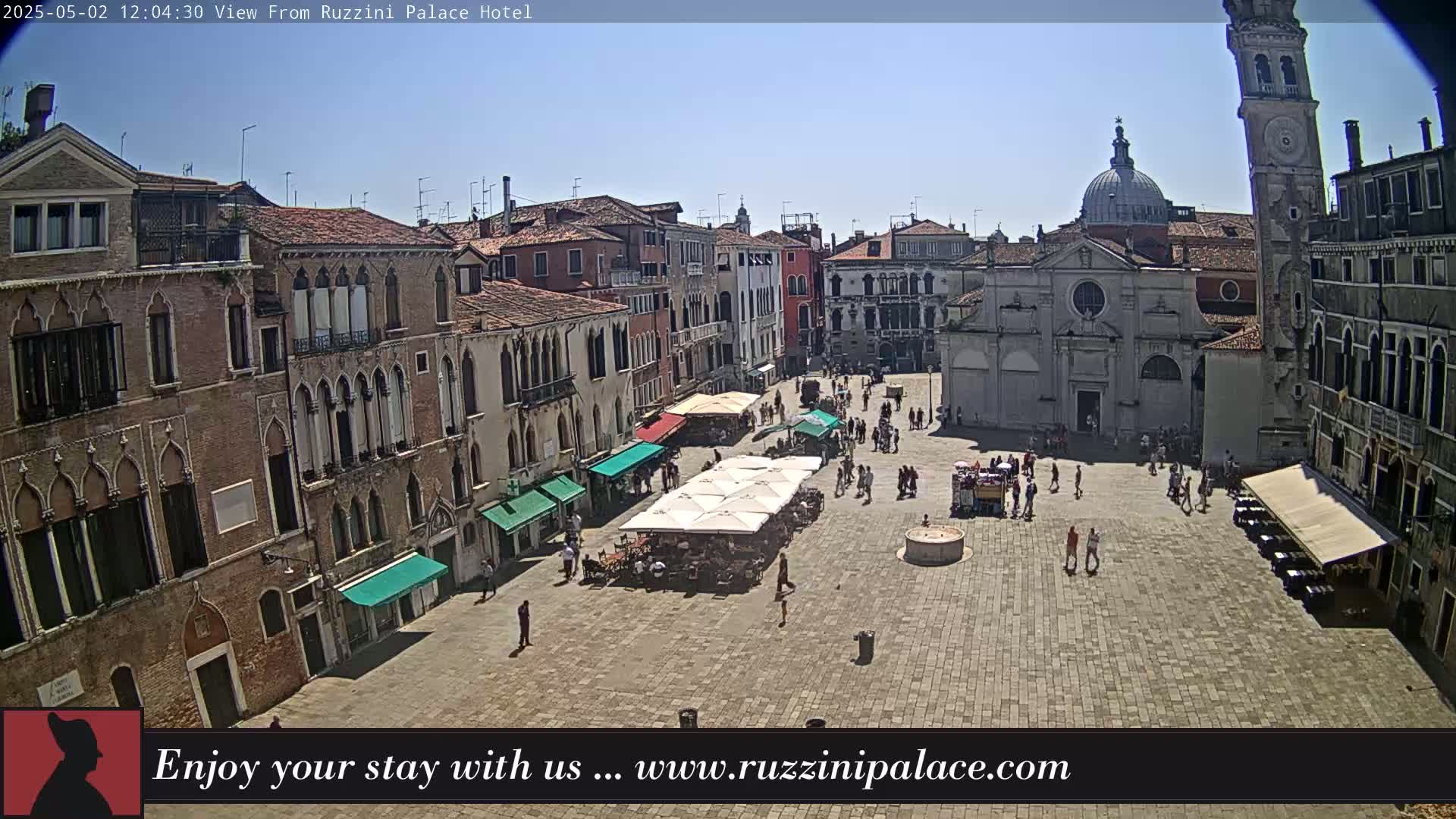A sunny Venetian square is filled with people and outdoor seating under umbrellas, surrounded by historic buildings including a church and tall tower.
