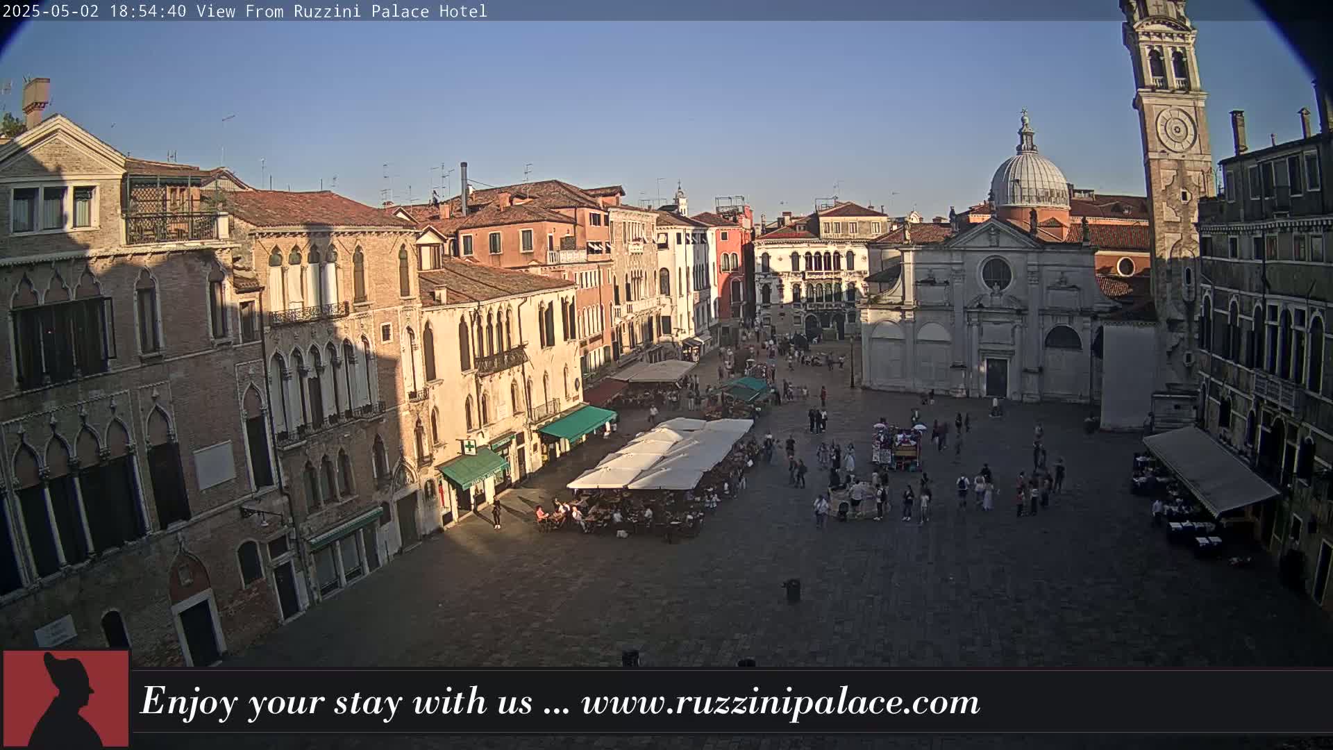 A sunlit Venetian square is filled with people and outdoor seating, surrounded by historic buildings under a clear sky.