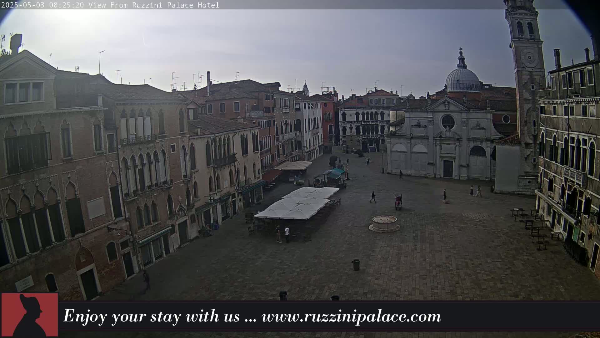 A mostly empty, stone plaza in Venice, under an overcast sky, is surrounded by old buildings, including a church and a tall tower.