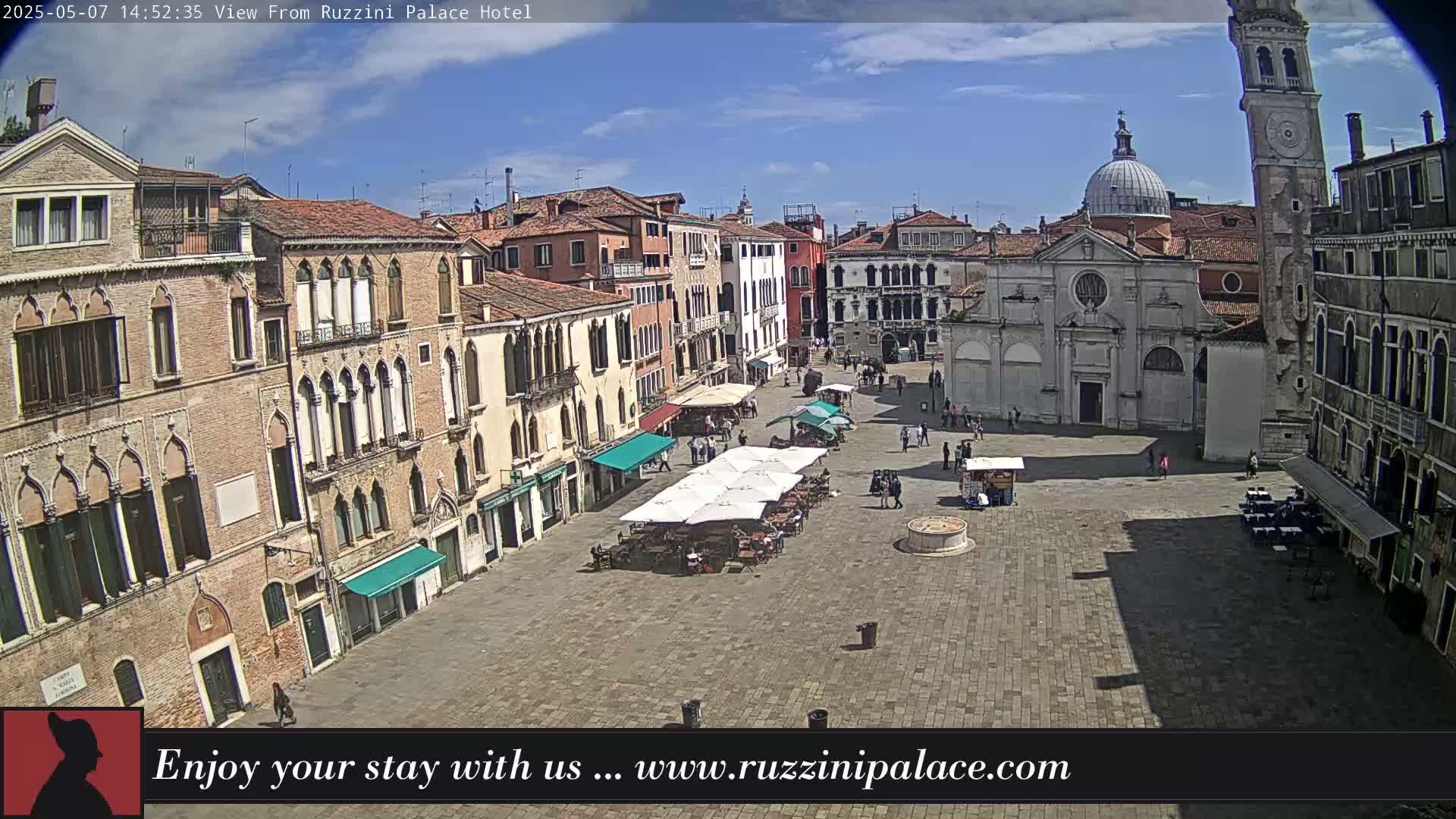 A sunny day reveals a large plaza in Venice, Italy, surrounded by historic buildings, with several people and outdoor seating areas visible.