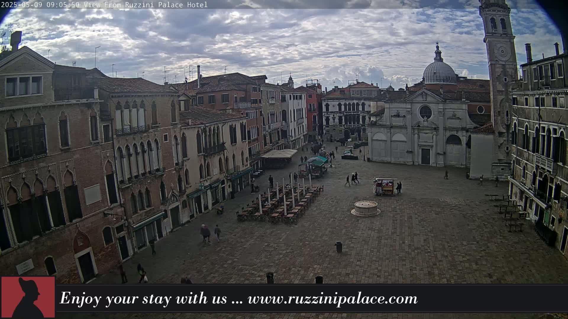A mostly cloudy day reveals a large, paved square in Venice, surrounded by historic buildings, with a few people and sparsely populated outdoor seating.