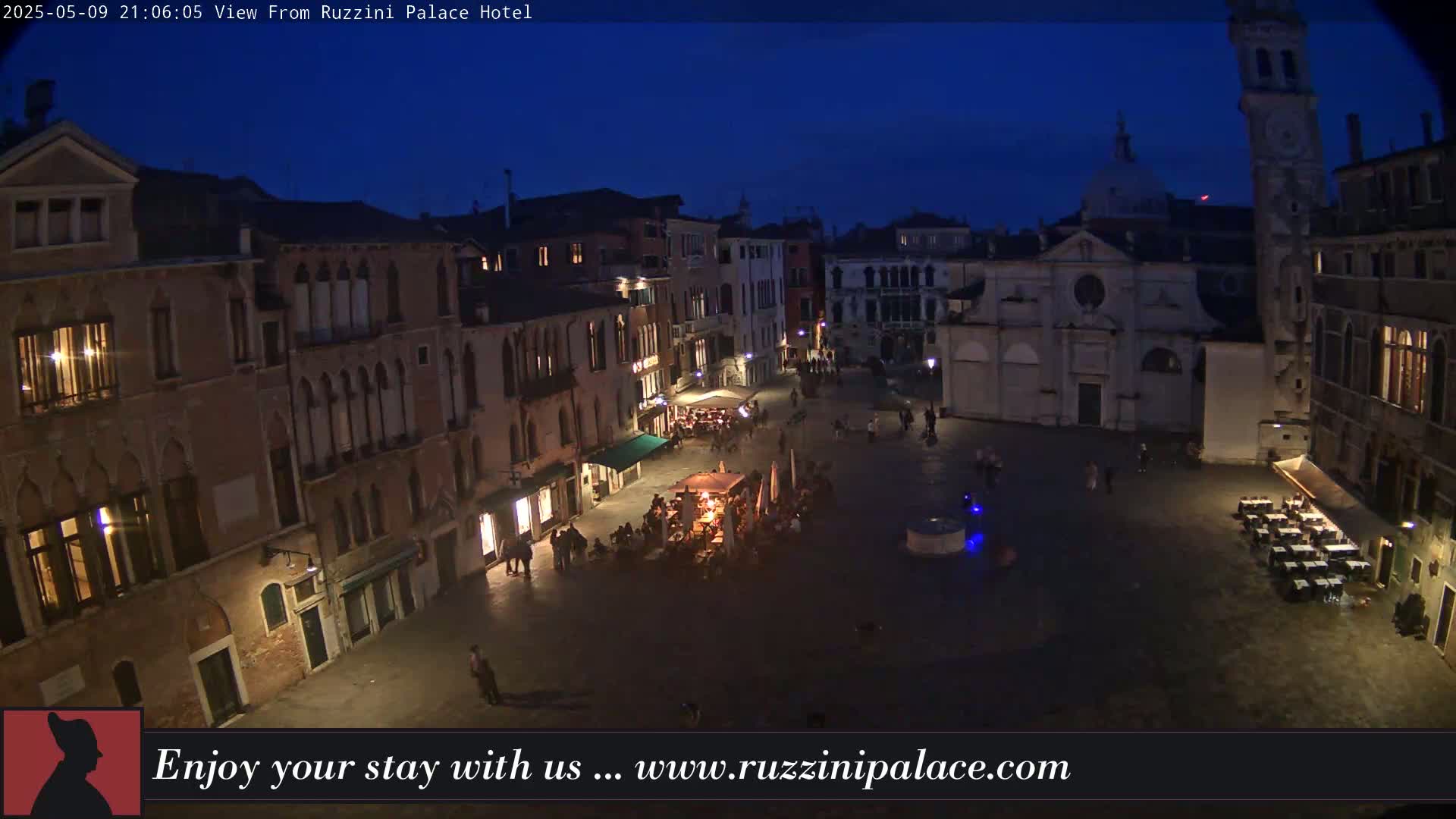 A nighttime aerial view of a dimly lit plaza in Venice, Italy, surrounded by old buildings, with a few people and outdoor seating areas visible.