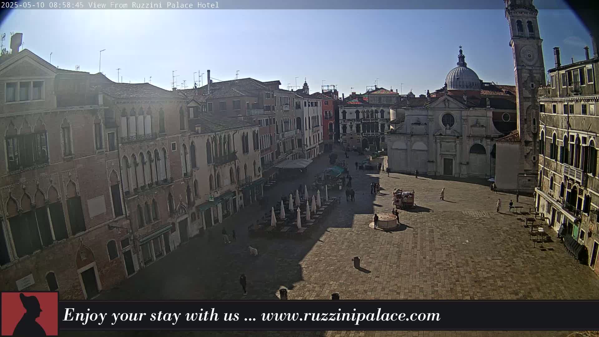 A sunny, paved square in Venice, Italy, is surrounded by several historic buildings, with some outdoor seating and a few people present.
