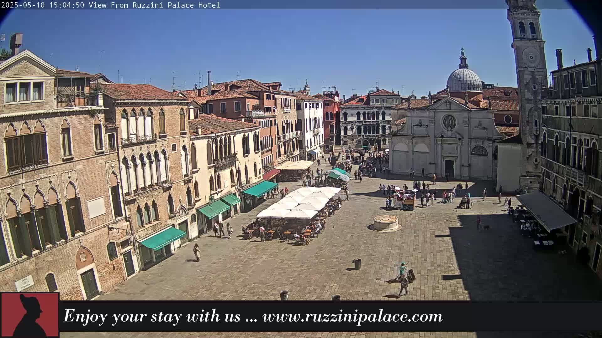 A sunny day reveals a large, paved square in Venice, surrounded by historic buildings, with people dining under outdoor umbrellas.