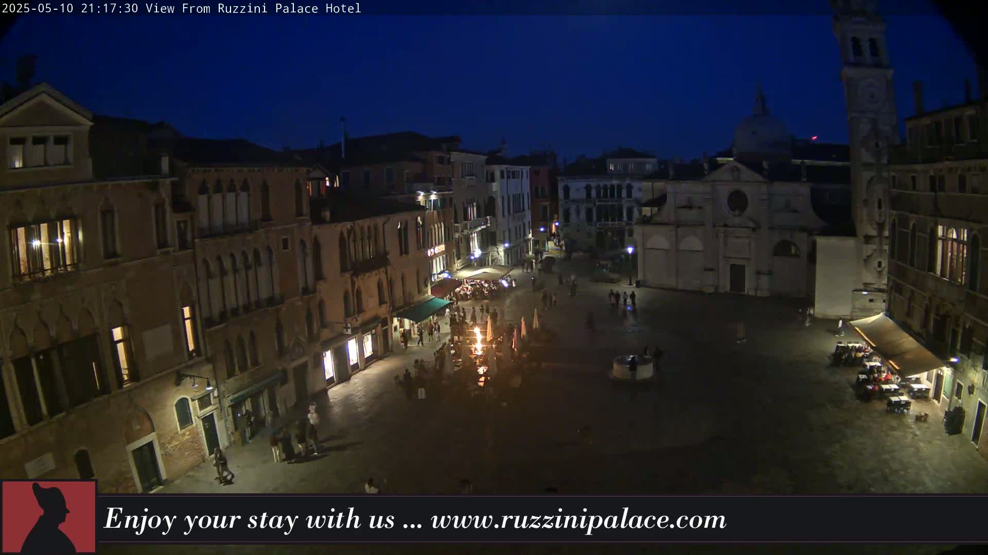 A nighttime aerial view of a dimly lit European plaza surrounded by old buildings, with a small fire burning in the center.