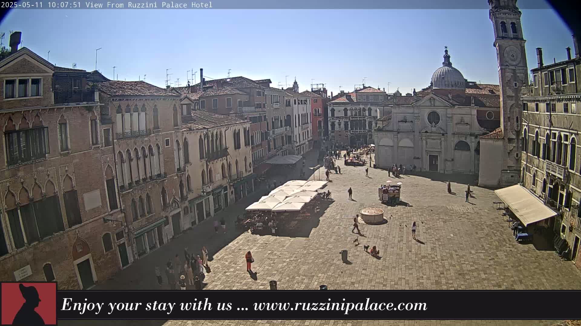 A sunny, paved square in Venice, Italy, is surrounded by several-story buildings, some with arched windows, and features outdoor seating under umbrellas.