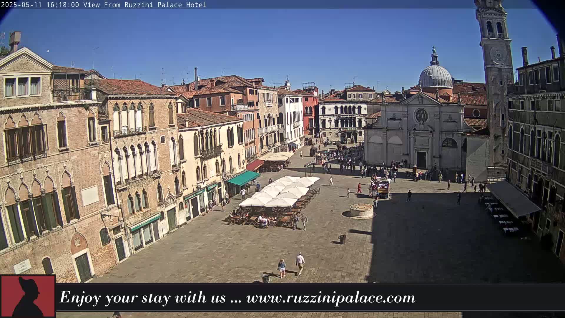 A sunny day reveals a large, cobblestone square in Venice, Italy, surrounded by old buildings, with several people and outdoor café seating visible.