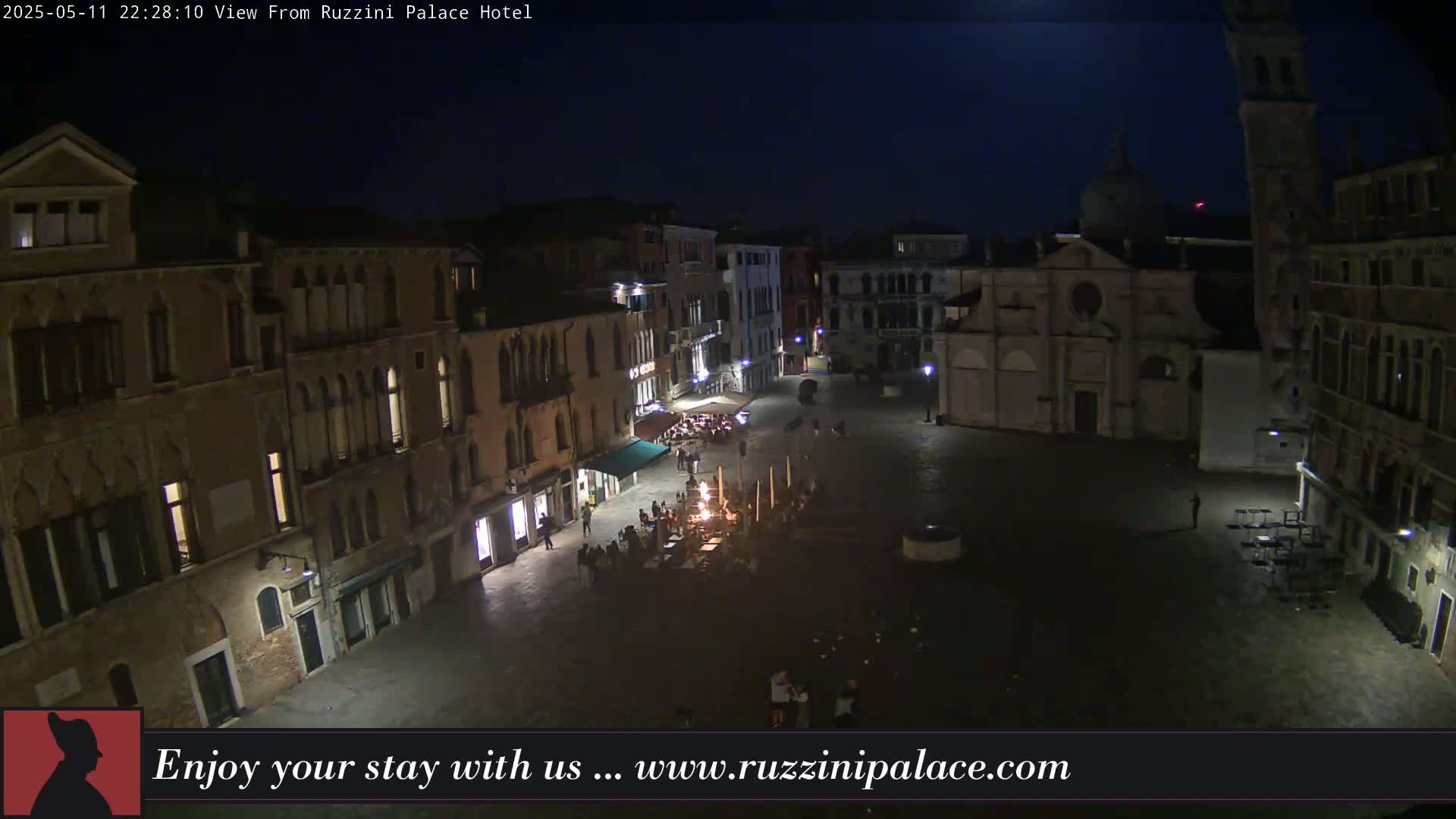 A nighttime aerial view shows a dimly lit plaza in Venice, surrounded by old buildings, with a few people gathered in the square.