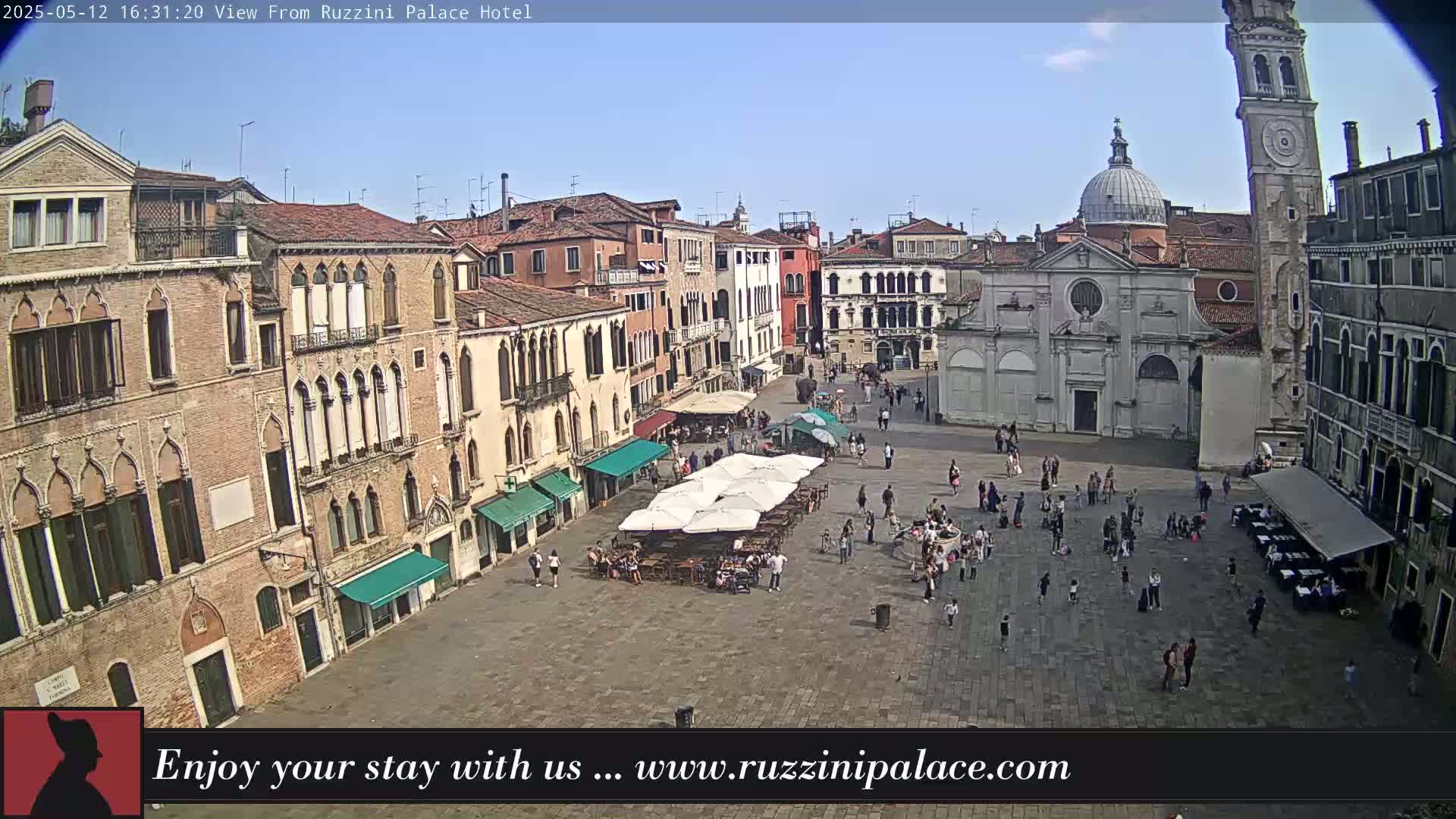 A sunny outdoor view of a large plaza in Venice, Italy, surrounded by historic buildings, with people strolling and sitting at outdoor cafes under umbrellas.