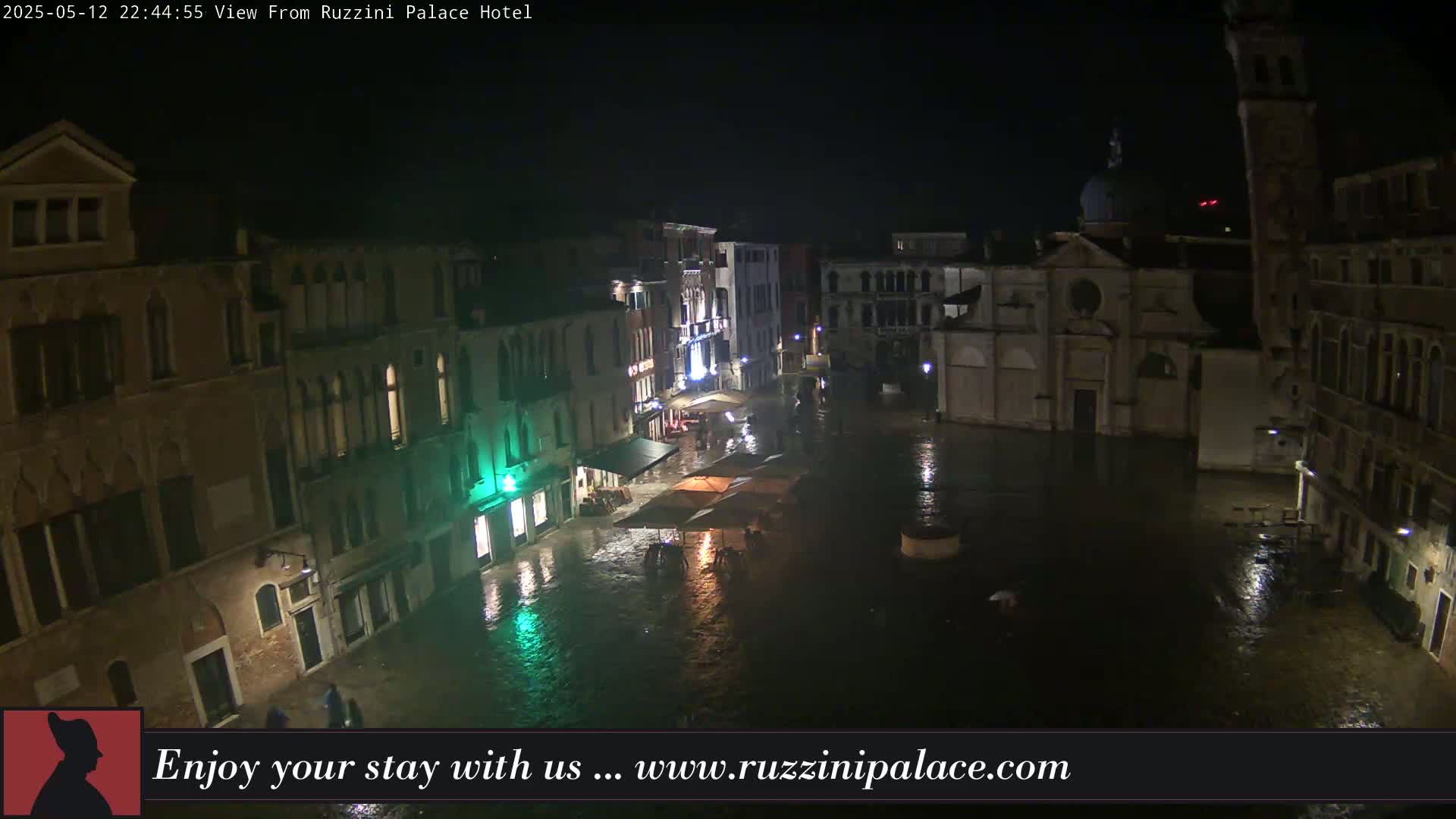A nighttime, rain-soaked Venetian square is seen from above, showing buildings and a few people.