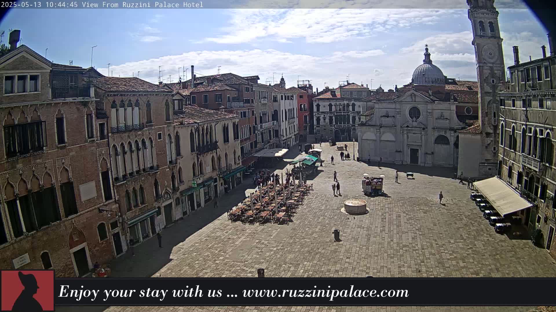 A sunny day in a large, paved European square surrounded by old brick buildings, featuring outdoor seating areas and a few people walking around.