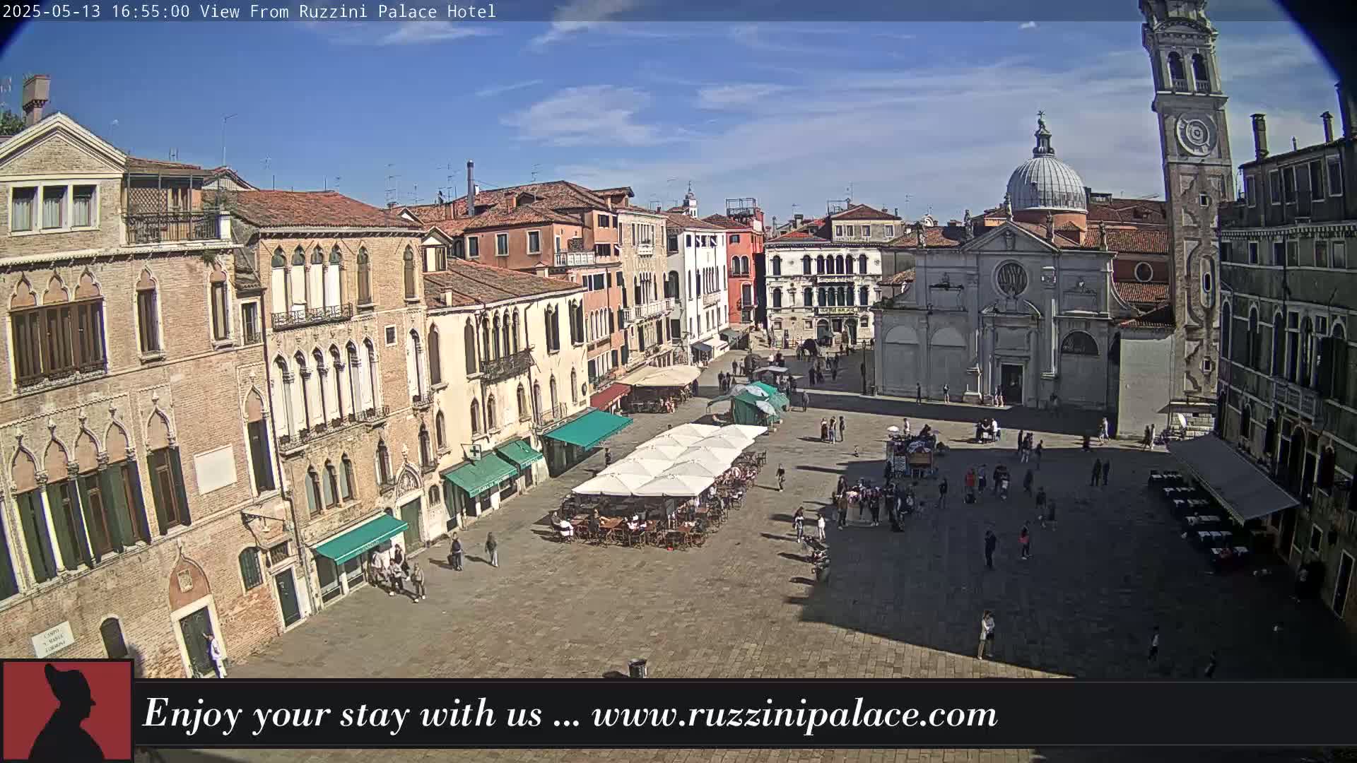 A sunny Venetian piazza with several buildings, a church, a tall bell tower, and outdoor seating under umbrellas.