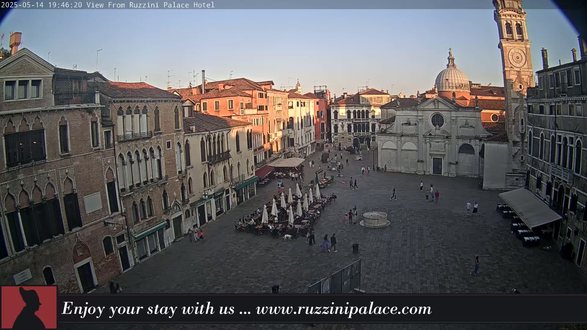 A large, paved square in Venice, Italy, is surrounded by old buildings under a clear, twilight sky; people are scattered throughout the square, some seated at outdoor cafes.