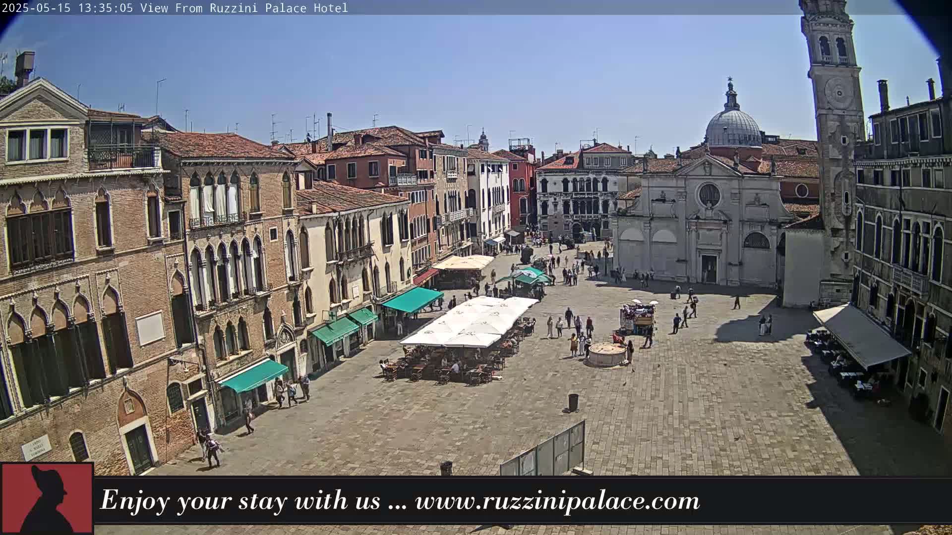 A sunny outdoor view of a large plaza in Venice, Italy, surrounded by historic buildings, with several people and outdoor seating areas.