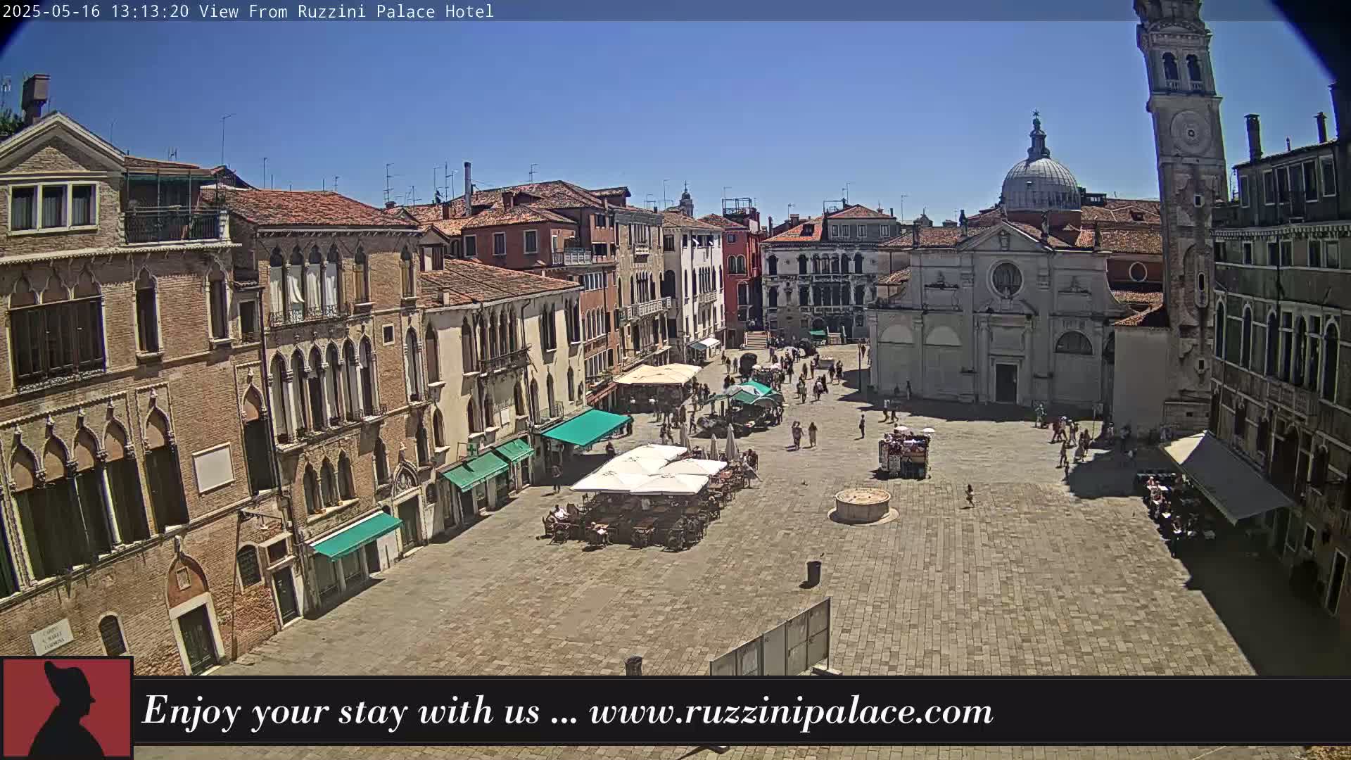 A sunny day in a large, paved town square surrounded by tall, old buildings, some with outdoor seating areas and a few people walking around.