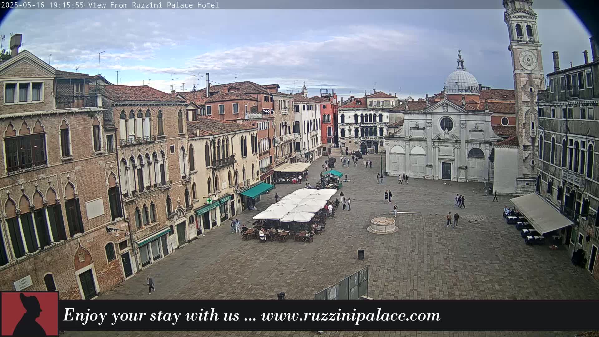 A mostly cloudy daytime view of a large, paved town square in Venice, Italy, surrounded by old buildings, with several people and outdoor seating areas under umbrellas.