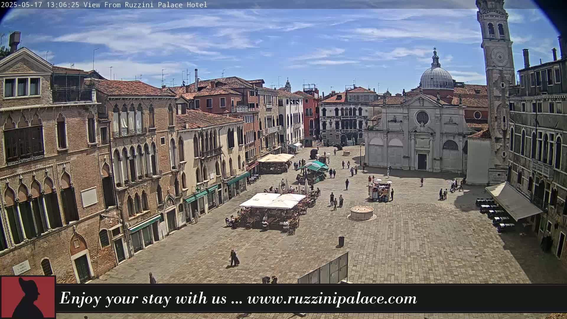 A sunny day shows a large, paved plaza in Venice, Italy, surrounded by historic buildings, with several people and outdoor seating areas.
