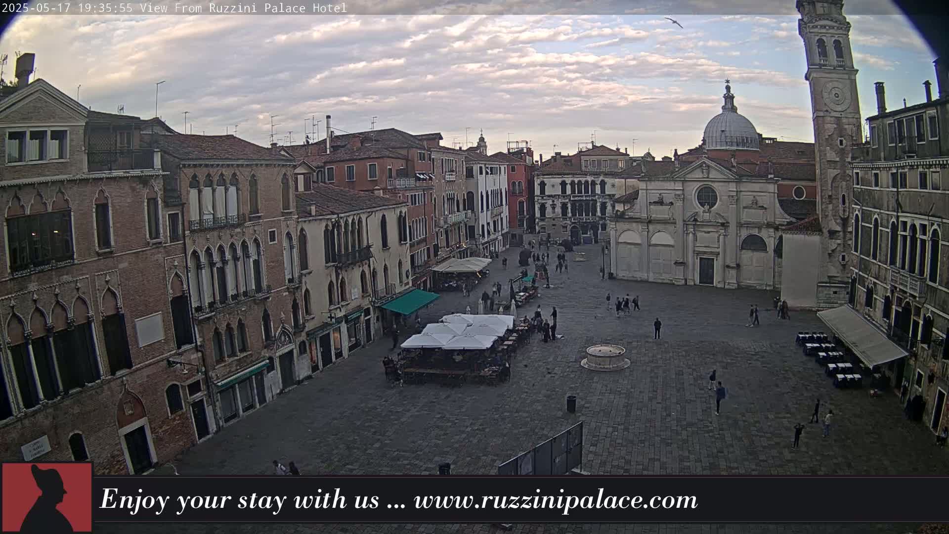 A mostly empty cobblestone square in Venice, Italy, is surrounded by old brick buildings under a partly cloudy evening sky.