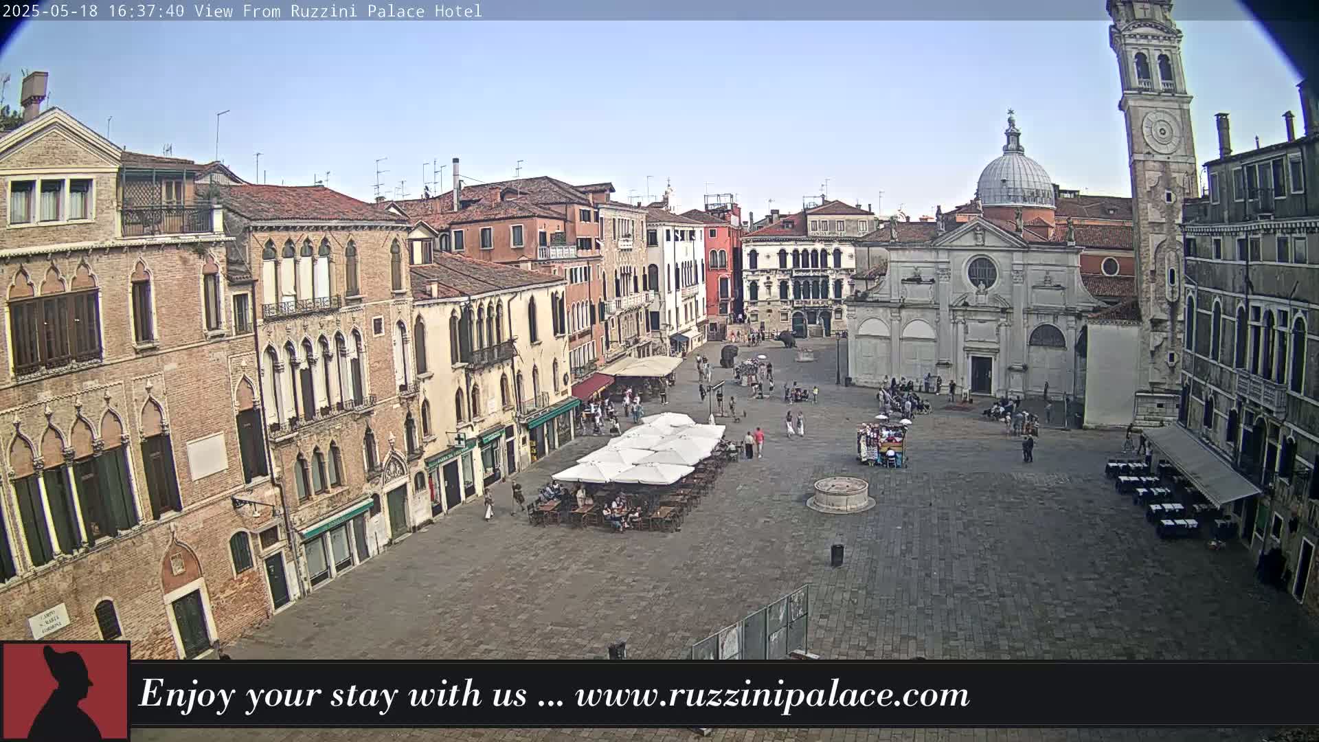 A large, paved town square in Venice, Italy, is surrounded by several-story buildings under a clear, mostly sunny sky;  people are scattered about, and several outdoor café umbrellas are set up.