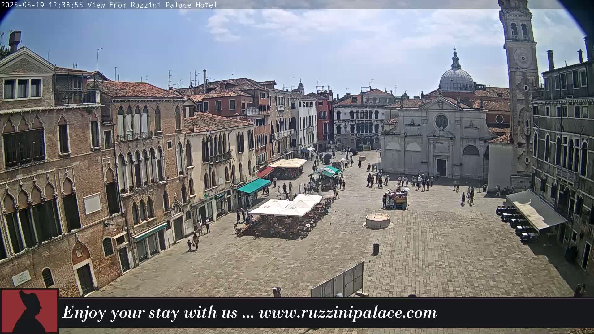 A sunny day reveals a large, paved town square in Venice, surrounded by historic buildings, with people strolling and some outdoor seating areas.